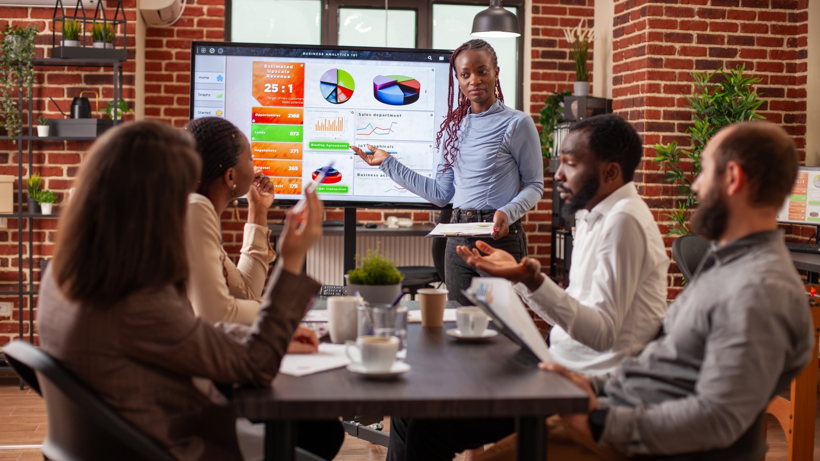 A woman presents data on a screen to a small team around a table in a brick-walled workspace